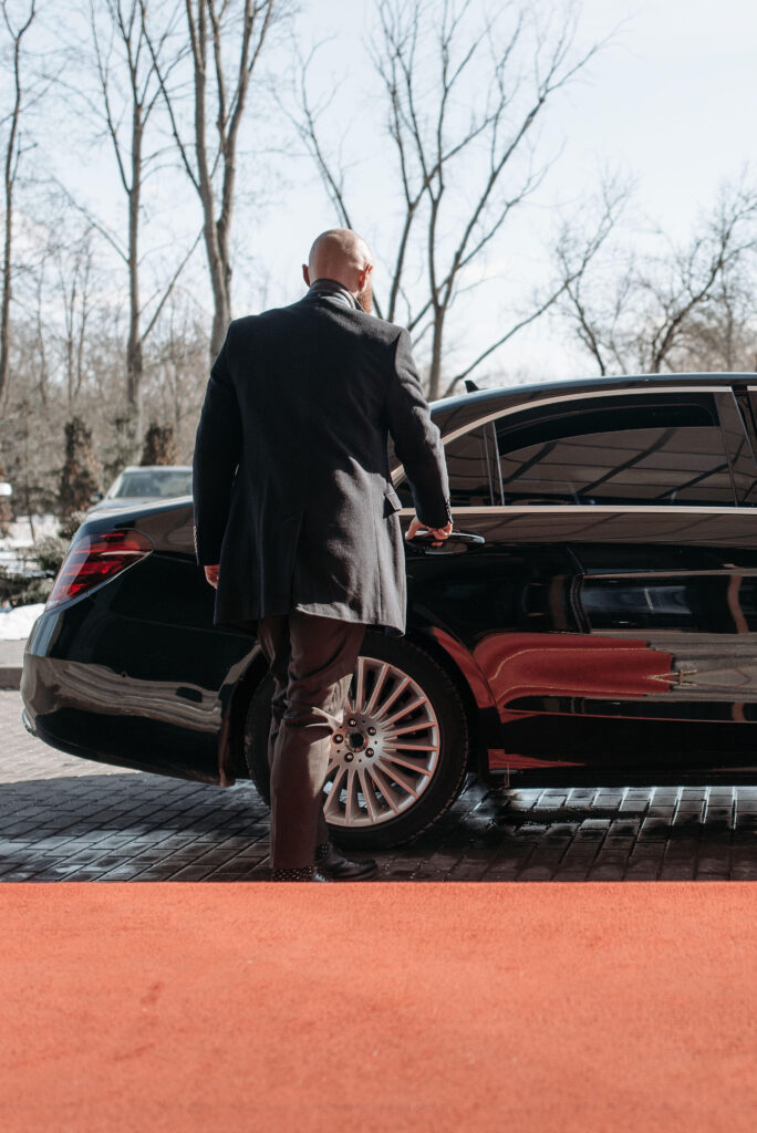 Man in formal attire approaching a black luxury car on a red carpet