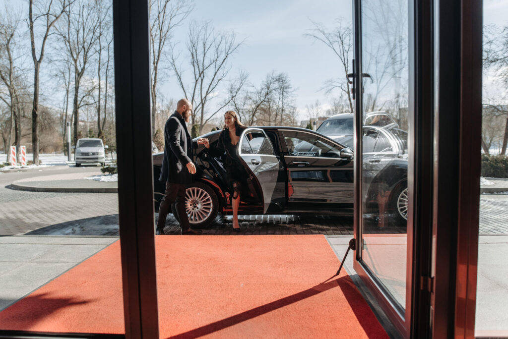 A woman exiting a luxury car onto a red carpet, with trees in the background.